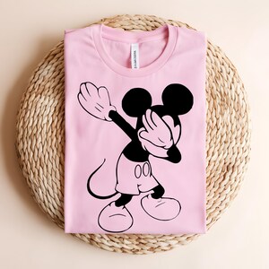 K&ouml;nnte beinhalten: Ein hellrosa T-Shirt mit kurzen &Auml;rmeln und einem schwarzen Grafikdruck der Zeichentrickfigur Mickey Mouse, die eine Dab-Pose ausf&uuml;hrt. Das T-Shirt hat einen Rundhalsausschnitt und ein sichtbares wei&szlig;es Markenetikett im Kragenbereich.