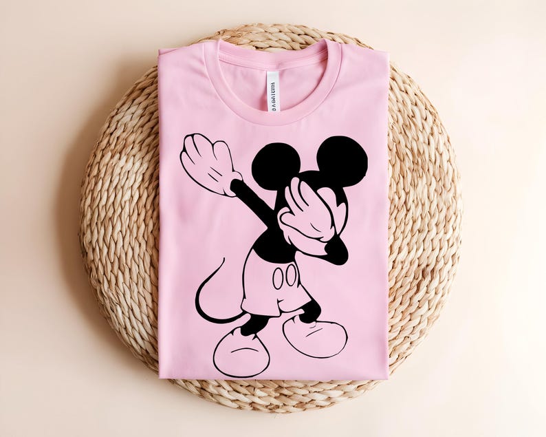 K&ouml;nnte beinhalten: Ein hellrosa T-Shirt mit kurzen &Auml;rmeln und einem schwarzen Grafikdruck der Zeichentrickfigur Mickey Mouse, die eine Dab-Pose ausf&uuml;hrt. Das T-Shirt hat einen Rundhalsausschnitt und ein sichtbares wei&szlig;es Markenetikett im Kragenbereich.