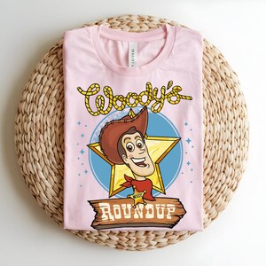 Puede incluir: Una camiseta rosa claro con un dise&ntilde;o de dibujos animados de Woody de Toy Story. El dise&ntilde;o incluye la cara de Woody, una estrella y las palabras "Woody's Roundup" en una fuente divertida. La camiseta est&aacute; doblada sobre una superficie tejida.