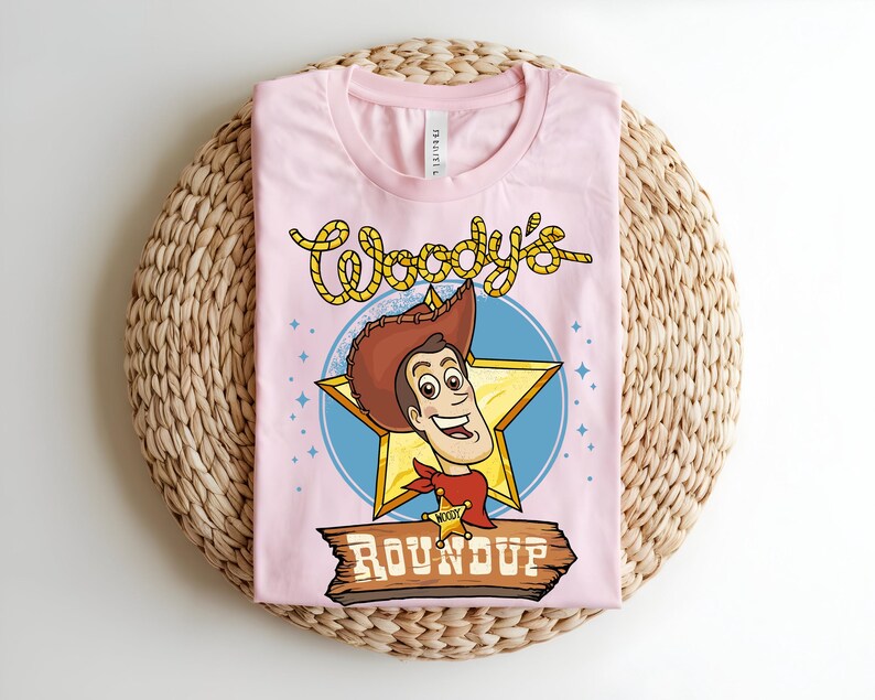 Puede incluir: Una camiseta rosa claro con un dise&ntilde;o de dibujos animados de Woody de Toy Story. El dise&ntilde;o incluye la cara de Woody, una estrella y las palabras "Woody's Roundup" en una fuente divertida. La camiseta est&aacute; doblada sobre una superficie tejida.