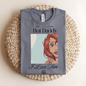 May include: A slate blue t-shirt featuring a graphic print of a red-haired character. The text reads "EST 1989", "But Daddy", and "I Love Him" in black lettering, with a small seashell icon centered at the bottom of the graphic design.