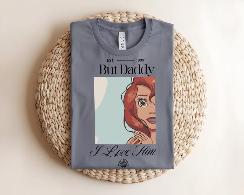 May include: A slate blue t-shirt featuring a graphic print of a red-haired character. The text reads "EST 1989", "But Daddy", and "I Love Him" in black lettering, with a small seashell icon centered at the bottom of the graphic design.