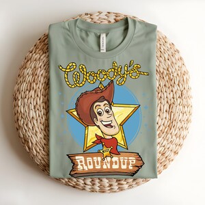 Puede incluir: Una camiseta verde salvia con una ilustraci&oacute;n de dibujos animados de Woody de Toy Story. El dise&ntilde;o incluye la cara de Woody, una estrella dorada y el texto "Woody's Roundup". La camiseta est&aacute; doblada sobre una estera tejida.