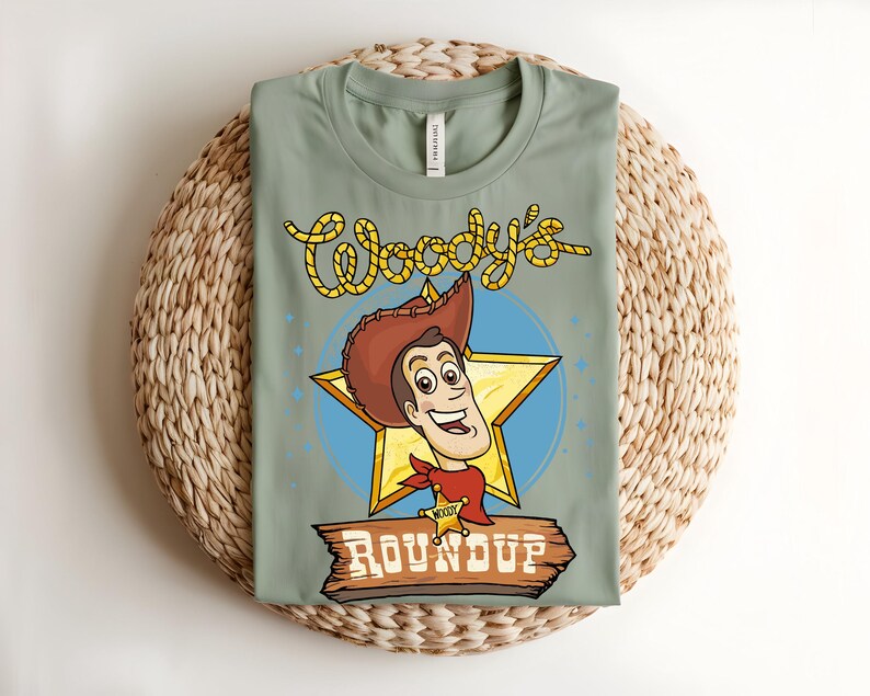 Puede incluir: Una camiseta verde salvia con una ilustraci&oacute;n de dibujos animados de Woody de Toy Story. El dise&ntilde;o incluye la cara de Woody, una estrella dorada y el texto "Woody's Roundup". La camiseta est&aacute; doblada sobre una estera tejida.