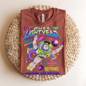 May include: A terracotta-coloured t-shirt with a graphic of Buzz Lightyear in a space scene. The design includes the words "Buzz Lightyear," "To Infinity and Beyond," and "Prepare for Ignition" in various colours. The shirt is displayed on a woven mat.