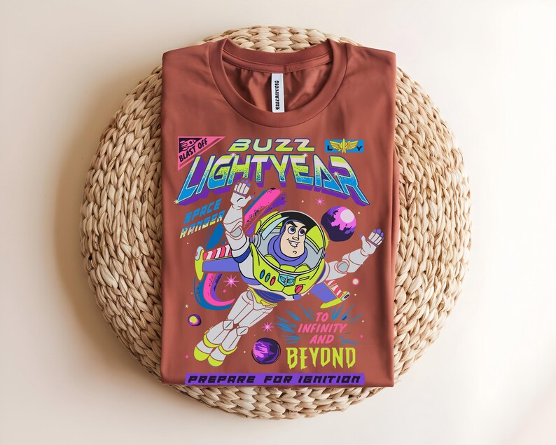 May include: A terracotta-coloured t-shirt with a graphic of Buzz Lightyear in a space scene. The design includes the words "Buzz Lightyear," "To Infinity and Beyond," and "Prepare for Ignition" in various colours. The shirt is displayed on a woven mat.