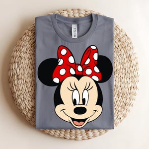Puede incluir: Una camiseta gris de cuello redondo con un estampado gr&aacute;fico de la cara de un rat&oacute;n de dibujos animados con grandes orejas negras y un lazo rojo con lunares blancos. La camiseta tiene una etiqueta en el cuello con el texto SSNA en letras negras.