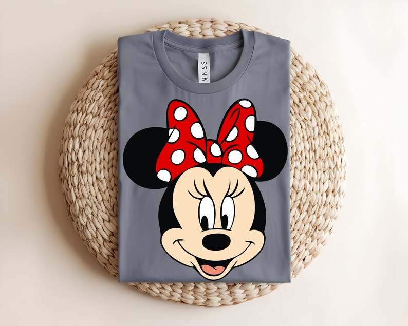 Puede incluir: Una camiseta gris de cuello redondo con un estampado gr&aacute;fico de la cara de un rat&oacute;n de dibujos animados con grandes orejas negras y un lazo rojo con lunares blancos. La camiseta tiene una etiqueta en el cuello con el texto SSNA en letras negras.