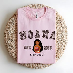 May include: A pale pink t-shirt featuring the word "MOANA" in a brown collegiate font. Below the word is a cartoon illustration of a woman and the text "EST. 2016 MOTUNUI". The shirt is folded on a woven, light brown surface.