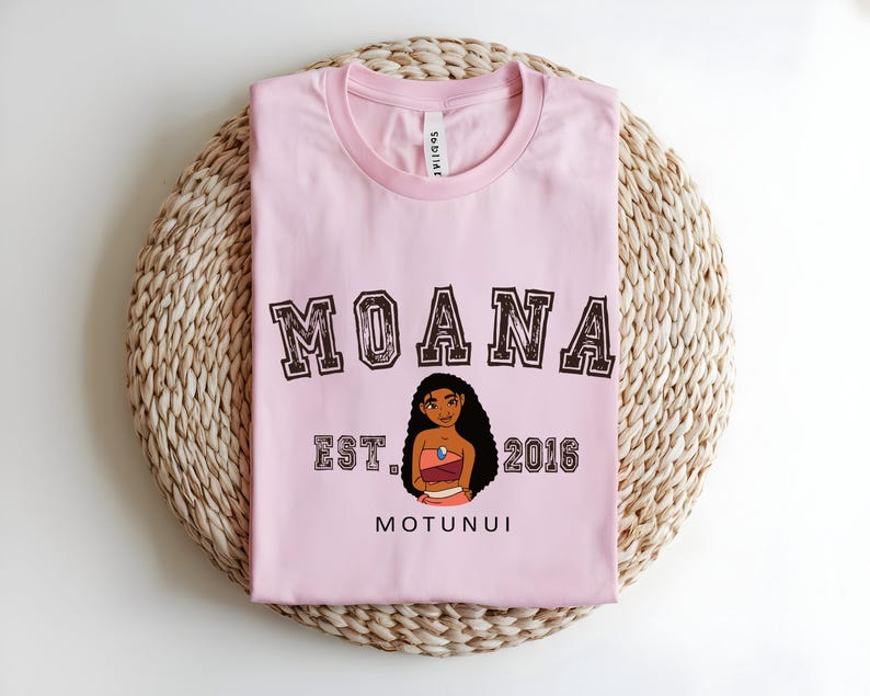 May include: A pale pink t-shirt featuring the word "MOANA" in a brown collegiate font. Below the word is a cartoon illustration of a woman and the text "EST. 2016 MOTUNUI". The shirt is folded on a woven, light brown surface.