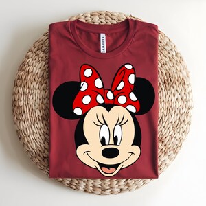 Puede incluir: Una camiseta de cuello redondo de color rojo oscuro con un gran estampado gr&aacute;fico de Minnie Mouse en la parte delantera. El personaje tiene la cabeza negra, la cara de color crema y un lazo rojo brillante con lunares blancos entre las orejas.