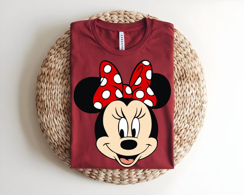 Puede incluir: Una camiseta de cuello redondo de color rojo oscuro con un gran estampado gr&aacute;fico de Minnie Mouse en la parte delantera. El personaje tiene la cabeza negra, la cara de color crema y un lazo rojo brillante con lunares blancos entre las orejas.
