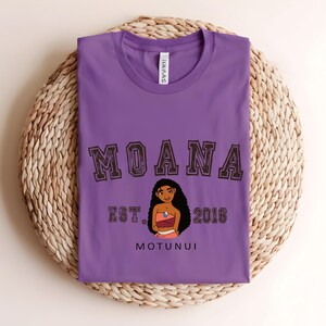 May include: A purple t-shirt featuring the word "MOANA" in brown block letters, a cartoon illustration of a woman, and the text "EST. 2016 MOTUNUI". The shirt is folded on a woven, circular surface.