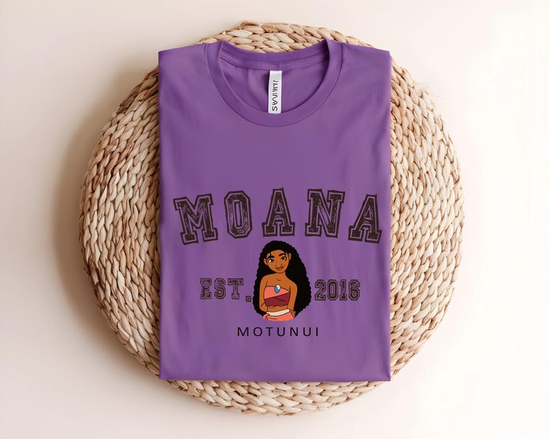 May include: A purple t-shirt featuring the word "MOANA" in brown block letters, a cartoon illustration of a woman, and the text "EST. 2016 MOTUNUI". The shirt is folded on a woven, circular surface.