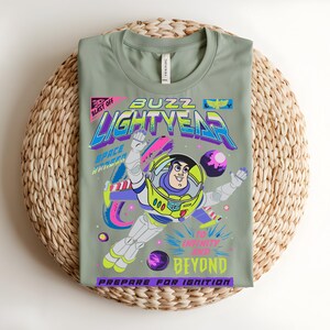 May include: Sage green t-shirt with a colourful graphic of Buzz Lightyear. The design includes the words "Buzz Lightyear", "To Infinity and Beyond", and "Prepare for Ignition". The t-shirt is folded on a woven mat, a popular home decor item.