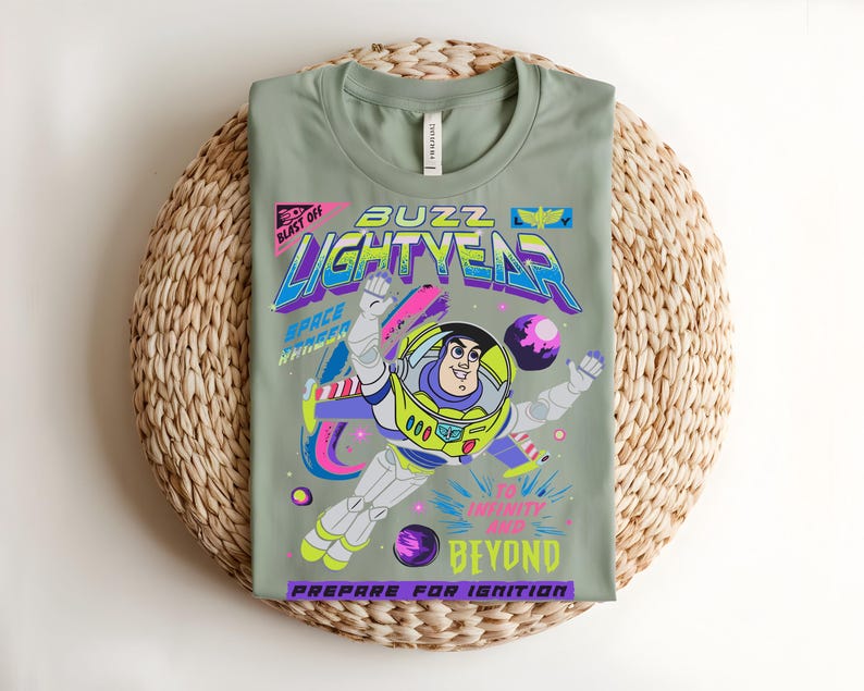 May include: Sage green t-shirt with a colourful graphic of Buzz Lightyear. The design includes the words "Buzz Lightyear", "To Infinity and Beyond", and "Prepare for Ignition". The t-shirt is folded on a woven mat, a popular home decor item.