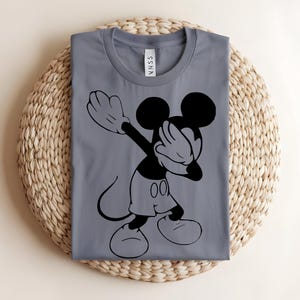 K&ouml;nnte beinhalten: Ein graues Kurzarm-T-Shirt mit einem schwarzen Grafikdruck einer Zeichentrick-Mausfigur, die eine Dabbing-Pose ausf&uuml;hrt. Das T-Shirt hat einen Rundhalsausschnitt und ein wei&szlig;es Innenetikett am Nacken mit dem Text SSNA in schwarzer Schrift.