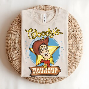 Puede incluir: Camiseta beige con una ilustraci&oacute;n de dibujos animados de un personaje de vaquero con sombrero marr&oacute;n, pa&ntilde;uelo rojo y placa de sheriff. La camiseta tiene el texto "Woody's Roundup" en amarillo y marr&oacute;n, sobre una estrella azul y un letrero de madera.
