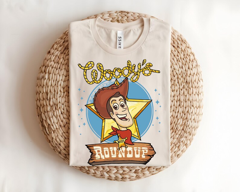 Puede incluir: Camiseta beige con una ilustraci&oacute;n de dibujos animados de un personaje de vaquero con sombrero marr&oacute;n, pa&ntilde;uelo rojo y placa de sheriff. La camiseta tiene el texto "Woody's Roundup" en amarillo y marr&oacute;n, sobre una estrella azul y un letrero de madera.