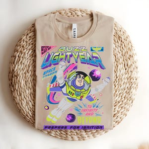 May include: Beige t-shirt with a vibrant Buzz Lightyear graphic. The design includes the words "BUZZ LIGHTYEAR", "TO INFINITY AND BEYOND", and "PREPARE FOR IGNITION". The shirt is folded on a woven mat.