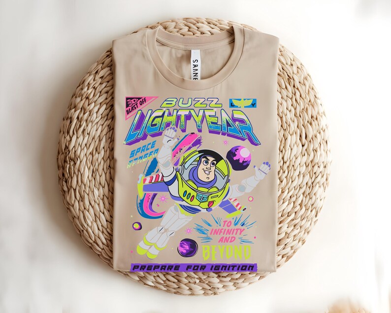 May include: Beige t-shirt with a vibrant Buzz Lightyear graphic. The design includes the words "BUZZ LIGHTYEAR", "TO INFINITY AND BEYOND", and "PREPARE FOR IGNITION". The shirt is folded on a woven mat.