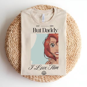 May include: A beige t-shirt featuring a graphic of a red-haired character with the text EST 1989, But Daddy, and I Love Him, along with a small seashell icon at the bottom. The shirt has a crew neckline and is displayed in a folded, flat layout.