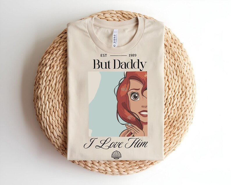 May include: A beige t-shirt featuring a graphic of a red-haired character with the text EST 1989, But Daddy, and I Love Him, along with a small seashell icon at the bottom. The shirt has a crew neckline and is displayed in a folded, flat layout.