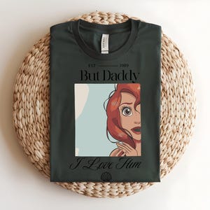 May include: A dark green t-shirt featuring a graphic print of a red-haired character. The text reads "EST 1989 But Daddy I Love Him" with a small seashell icon at the bottom. The shirt has a crew neckline and a standard short-sleeve design.