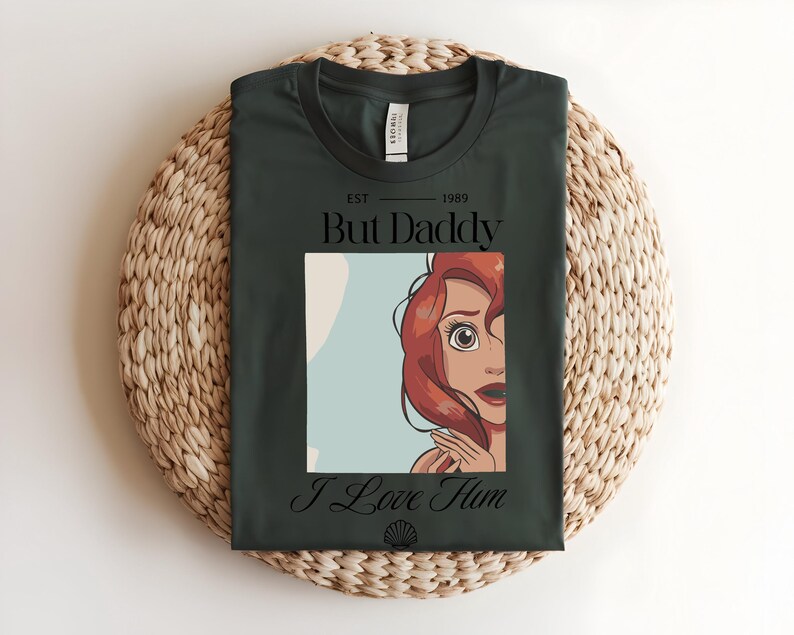 May include: A dark green t-shirt featuring a graphic print of a red-haired character. The text reads "EST 1989 But Daddy I Love Him" with a small seashell icon at the bottom. The shirt has a crew neckline and a standard short-sleeve design.