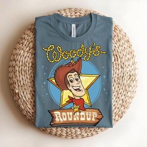 Puede incluir: Una camiseta azul con un gr&aacute;fico de dibujos animados de Woody de Toy Story. El dise&ntilde;o incluye la cara de Woody, una estrella y el texto "Woody's Roundup". La camiseta est&aacute; doblada y colocada sobre una superficie tejida.