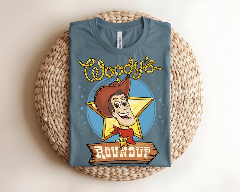 Puede incluir: Una camiseta azul con un gr&aacute;fico de dibujos animados de Woody de Toy Story. El dise&ntilde;o incluye la cara de Woody, una estrella y el texto "Woody's Roundup". La camiseta est&aacute; doblada y colocada sobre una superficie tejida.