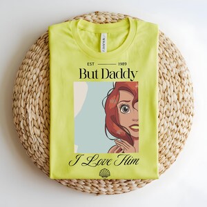 May include: A lime green t-shirt featuring a graphic of a red-haired character with large eyes. The text reads EST 1989 But Daddy I Love Him, with a small seashell icon at the bottom. The shirt has a crew neckline and a standard short-sleeve cut.