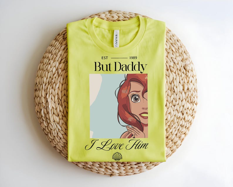 May include: A lime green t-shirt featuring a graphic of a red-haired character with large eyes. The text reads EST 1989 But Daddy I Love Him, with a small seashell icon at the bottom. The shirt has a crew neckline and a standard short-sleeve cut.