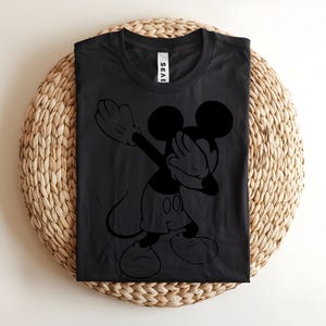 K&ouml;nnte beinhalten: Ein schwarzes Kurzarm-T-Shirt mit einer schwarzen grafischen Umrisszeichnung der Figur Mickey Mouse, die eine Dab-Tanzbewegung ausf&uuml;hrt. Das Shirt hat einen Rundhalsausschnitt und ein wei&szlig;es Markenetikett am inneren Kragenbereich auf dem dunklen Stoff.