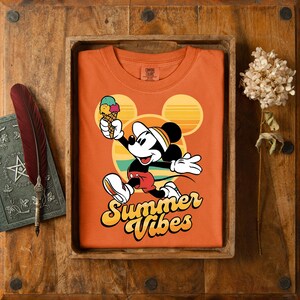 以下が含まれることがあります： ミッキーマウスがアイスクリームを持っているオレンジ色のTシャツ。 「Summer Vibes」の文字入り。 デザインには、黄色とオレンジ色の夕日とミッキーのシルエットが含まれています。 シャツは木製のトレイに、羽と本と一緒に表示されています。