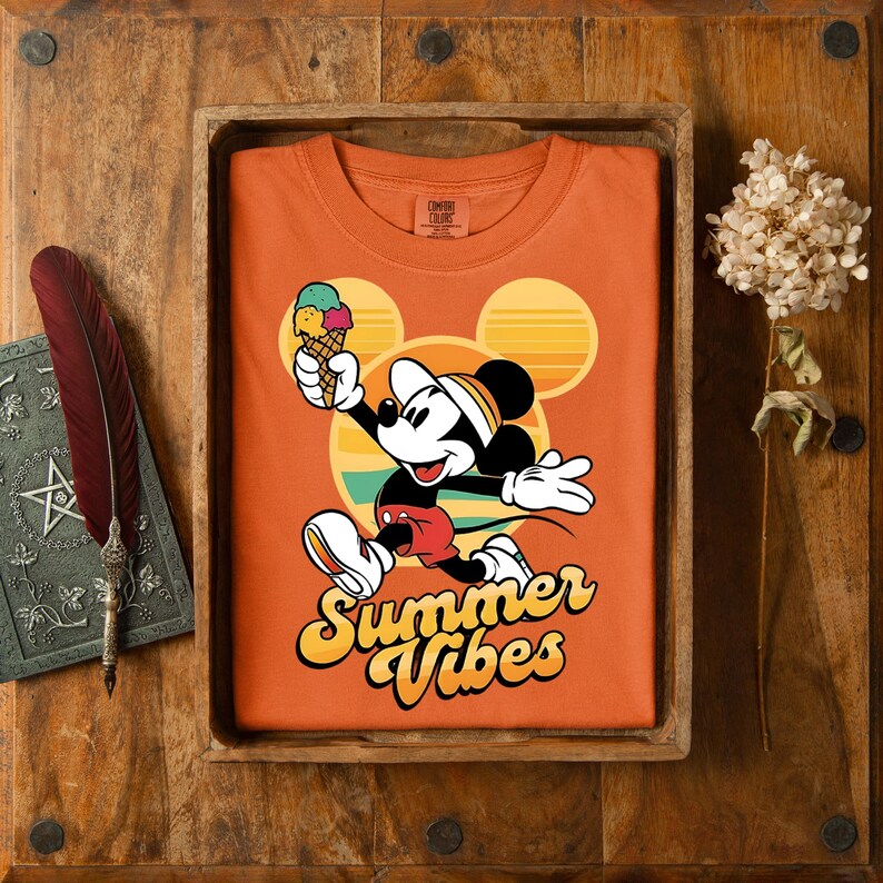 以下が含まれることがあります： ミッキーマウスがアイスクリームを持っているオレンジ色のTシャツ。 「Summer Vibes」の文字入り。 デザインには、黄色とオレンジ色の夕日とミッキーのシルエットが含まれています。 シャツは木製のトレイに、羽と本と一緒に表示されています。