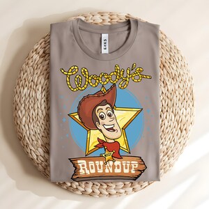 Puede incluir: Una camiseta gris con una imagen de dibujos animados de Woody de Toy Story. El dise&ntilde;o incluye la cara de Woody, una estrella dorada y el texto "Woody's Roundup" en una fuente de estilo occidental. La camiseta est&aacute; doblada sobre una estera tejida.