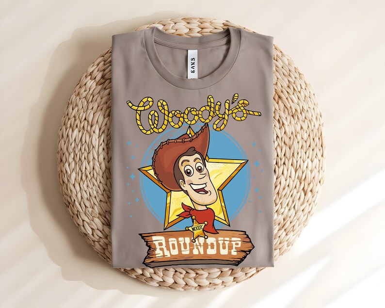 Puede incluir: Una camiseta gris con una imagen de dibujos animados de Woody de Toy Story. El dise&ntilde;o incluye la cara de Woody, una estrella dorada y el texto "Woody's Roundup" en una fuente de estilo occidental. La camiseta est&aacute; doblada sobre una estera tejida.