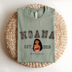 May include: Sage green t-shirt with the word "MOANA" in a collegiate font above a character illustration. Below the illustration are the words "EST. 2016" and "MOTUNUI". The shirt is folded on a woven mat.