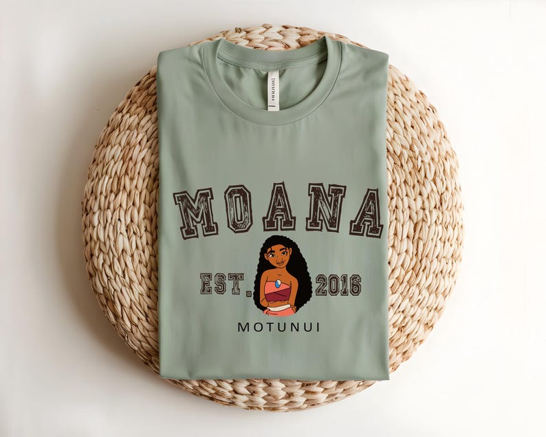 May include: Sage green t-shirt with the word "MOANA" in a collegiate font above a character illustration. Below the illustration are the words "EST. 2016" and "MOTUNUI". The shirt is folded on a woven mat.