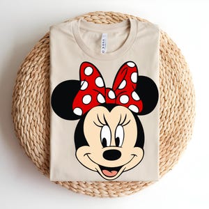 Puede incluir: Una camiseta de manga corta de color beige con un gran estampado gr&aacute;fico de Minnie Mouse en la parte delantera. El personaje tiene la cabeza negra, la cara color crema y un lazo rojo brillante con lunares blancos. La camiseta tiene cuello redondo.