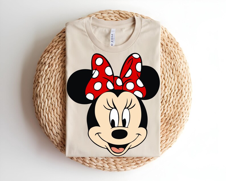 Puede incluir: Una camiseta de manga corta de color beige con un gran estampado gr&aacute;fico de Minnie Mouse en la parte delantera. El personaje tiene la cabeza negra, la cara color crema y un lazo rojo brillante con lunares blancos. La camiseta tiene cuello redondo.