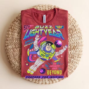 May include: A maroon t-shirt with a vibrant graphic of Buzz Lightyear from Toy Story. The design includes the words "Buzz Lightyear," "Space Ranger," and "To Infinity and Beyond." The shirt is folded on a woven, circular mat.