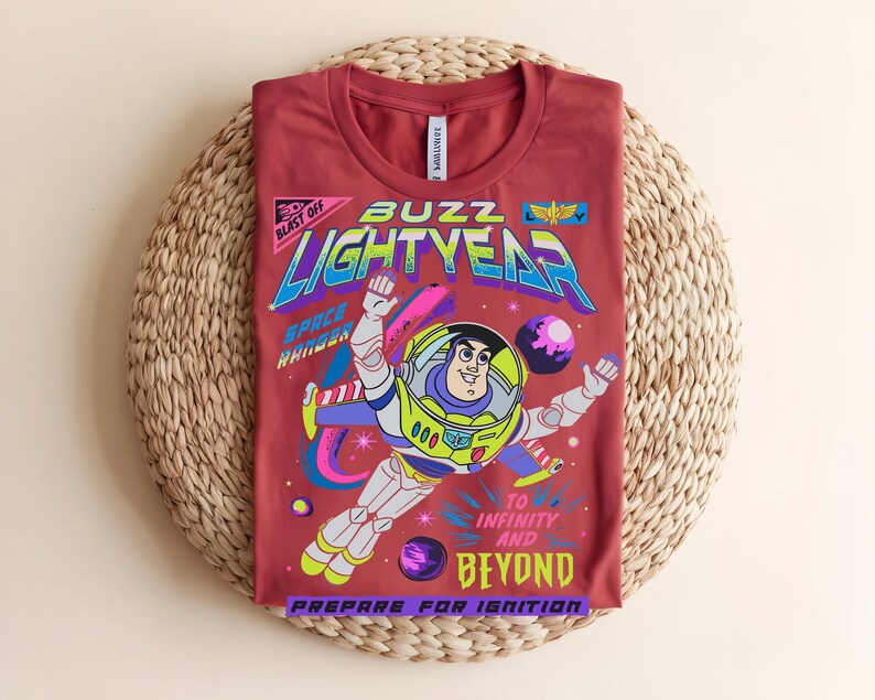 May include: A maroon t-shirt with a vibrant graphic of Buzz Lightyear from Toy Story. The design includes the words "Buzz Lightyear," "Space Ranger," and "To Infinity and Beyond." The shirt is folded on a woven, circular mat.