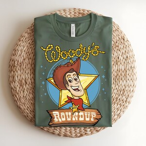Puede incluir: Camiseta verde con una imagen de dibujos animados de Woody de Toy Story. El dise&ntilde;o incluye la cara de Woody dentro de una estrella dorada, un c&iacute;rculo azul y las palabras "Woody's Roundup" en una fuente de estilo occidental. La camiseta se muestra sobre una estera tejida.