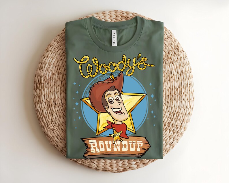 Puede incluir: Camiseta verde con una imagen de dibujos animados de Woody de Toy Story. El dise&ntilde;o incluye la cara de Woody dentro de una estrella dorada, un c&iacute;rculo azul y las palabras "Woody's Roundup" en una fuente de estilo occidental. La camiseta se muestra sobre una estera tejida.