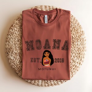 May include: A rust-coloured t-shirt with the word "MOANA" in a collegiate font above a character illustration. Below the illustration are the words "EST. 2016" and "MOTUNUI". The shirt is displayed on a woven, circular placemat.