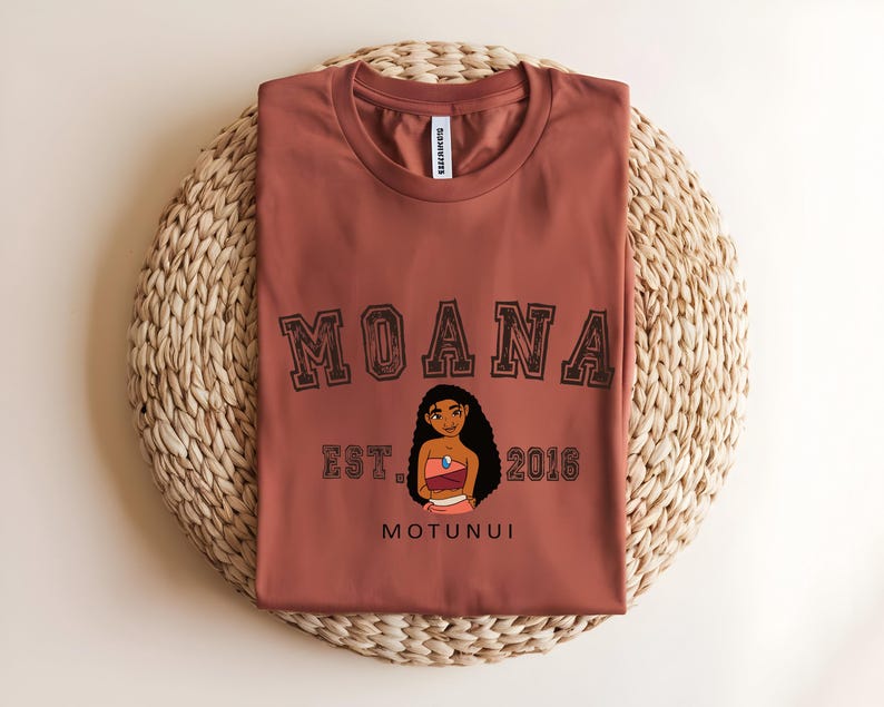 May include: A rust-coloured t-shirt with the word "MOANA" in a collegiate font above a character illustration. Below the illustration are the words "EST. 2016" and "MOTUNUI". The shirt is displayed on a woven, circular placemat.