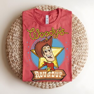 Puede incluir: Una camiseta roja con una ilustraci&oacute;n de dibujos animados de Woody de Toy Story. El dise&ntilde;o incluye la cara de Woody, una estrella y las palabras "Woody's Roundup" en una fuente de estilo occidental. La camiseta se muestra sobre una estera tejida.