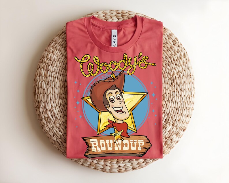 Puede incluir: Una camiseta roja con una ilustraci&oacute;n de dibujos animados de Woody de Toy Story. El dise&ntilde;o incluye la cara de Woody, una estrella y las palabras "Woody's Roundup" en una fuente de estilo occidental. La camiseta se muestra sobre una estera tejida.